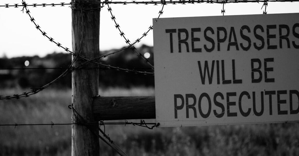 pexels-photo-11428401-11428401 Monochrome image of a barbed wire fence with a sign warning trespassers will be prosecuted.